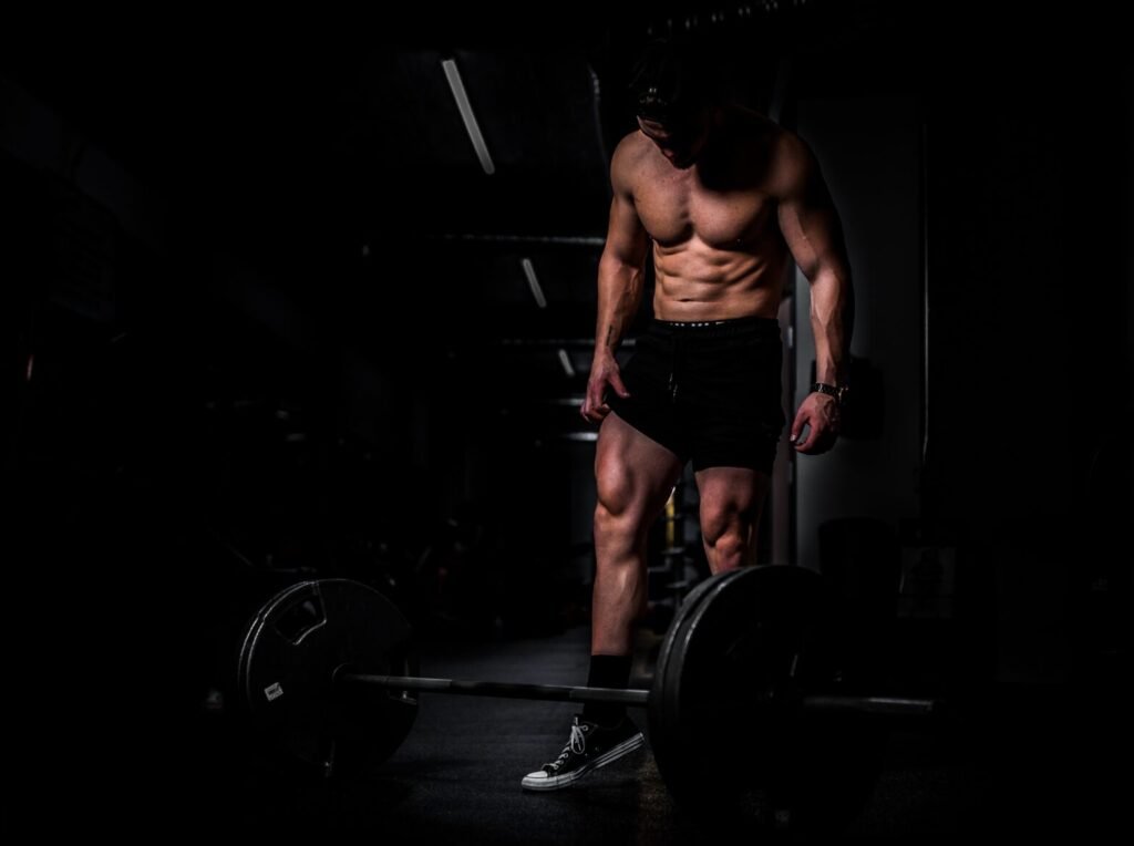 Man met ontbloot bovenlichaam staat voor een geladen barbell op de gymvloer in low-key belichting.